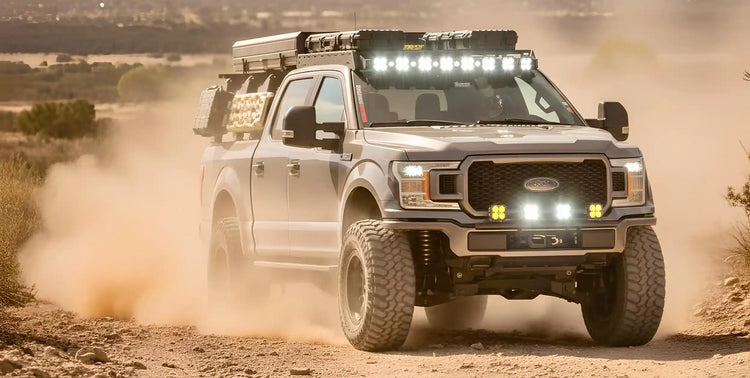 A heavily modified silver Ford pickup truck with large off-road tires, a roof rack, and multiple bright LED light bars driving on a dusty dirt road.
