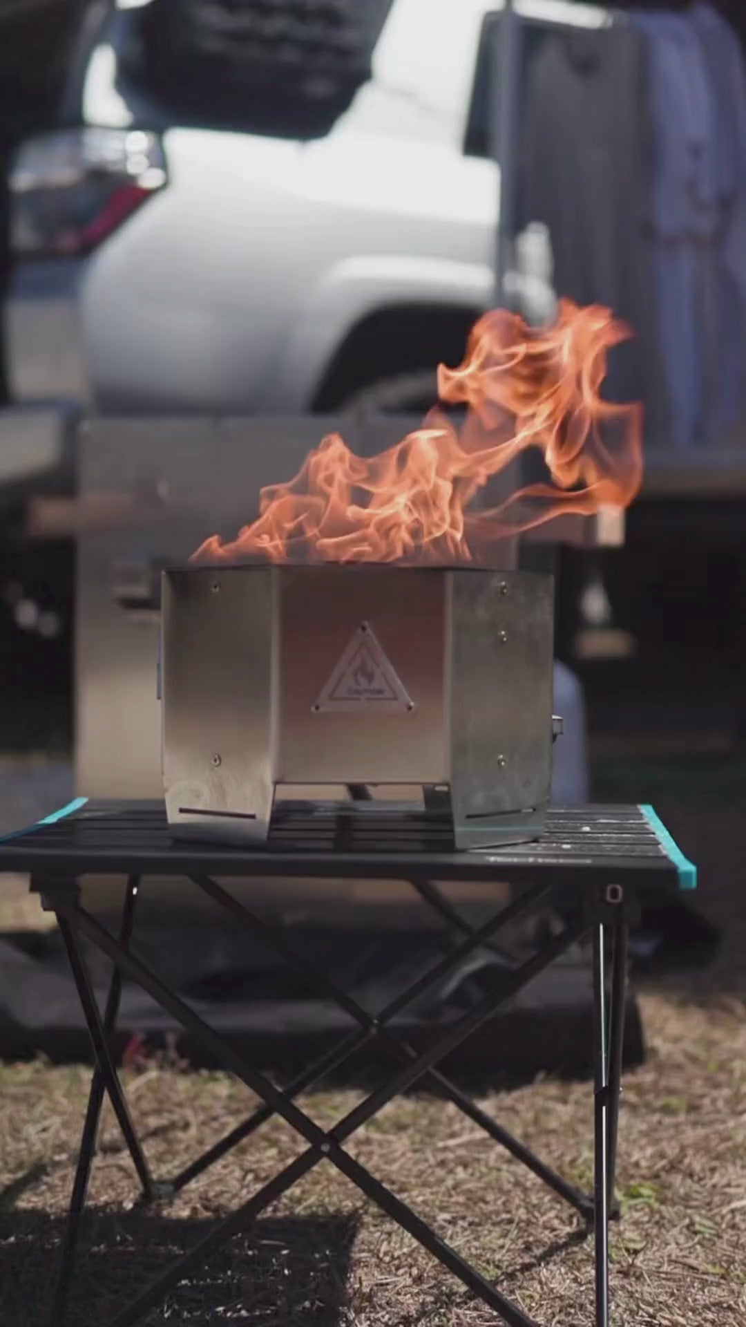 Close-up front view of a portable stainless steel propane fire pit burning brightly during a camping trip, showcasing its steady flame and outdoor design
