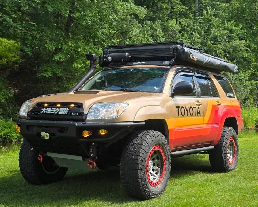 A customized Toyota 4Runner SUV parked on a grassy lawn with a forest background. The vehicle features a tan and orange paint job, a black roof rack with a top box, a front bumper guard, and off-road tires.