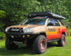 A customized Toyota 4Runner SUV parked on a grassy lawn with a forest background. The vehicle features a tan and orange paint job, a black roof rack with a top box, a front bumper guard, and off-road tires.