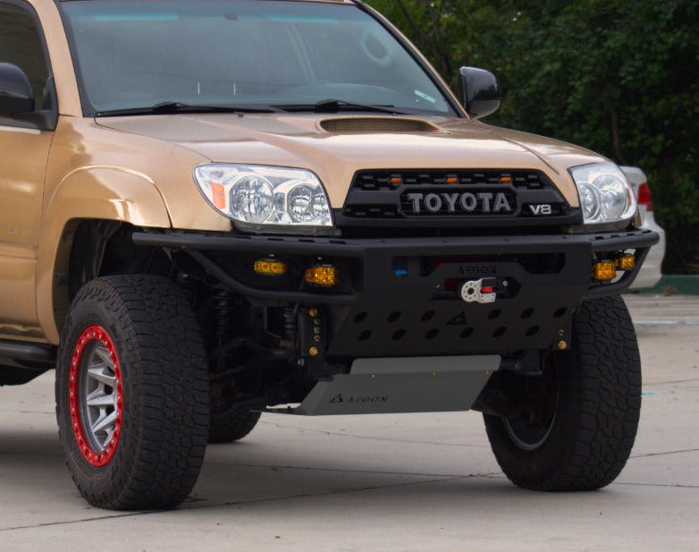 A tan Toyota 4Runner SUV with a black front bumper, roof rack, and off-road tires, parked outdoors on a paved surface with trees and a building in the background.
