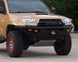 A tan Toyota 4Runner SUV with a black front bumper, roof rack, and off-road tires, parked outdoors on a paved surface with trees and a building in the background.