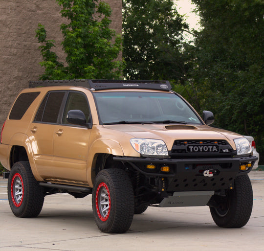 A tan Toyota 4Runner lifted SUV with a black front bumper, roof rack, and off-road tires, parked outdoors on a concrete surface with trees and a building in the background.
