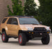 A tan Toyota 4Runner lifted SUV with a black front bumper, roof rack, and off-road tires, parked outdoors on a concrete surface with trees and a building in the background.