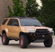 A tan Toyota 4Runner lifted SUV with a black front bumper, roof rack, and off-road tires, parked outdoors on a concrete surface with trees and a building in the background.