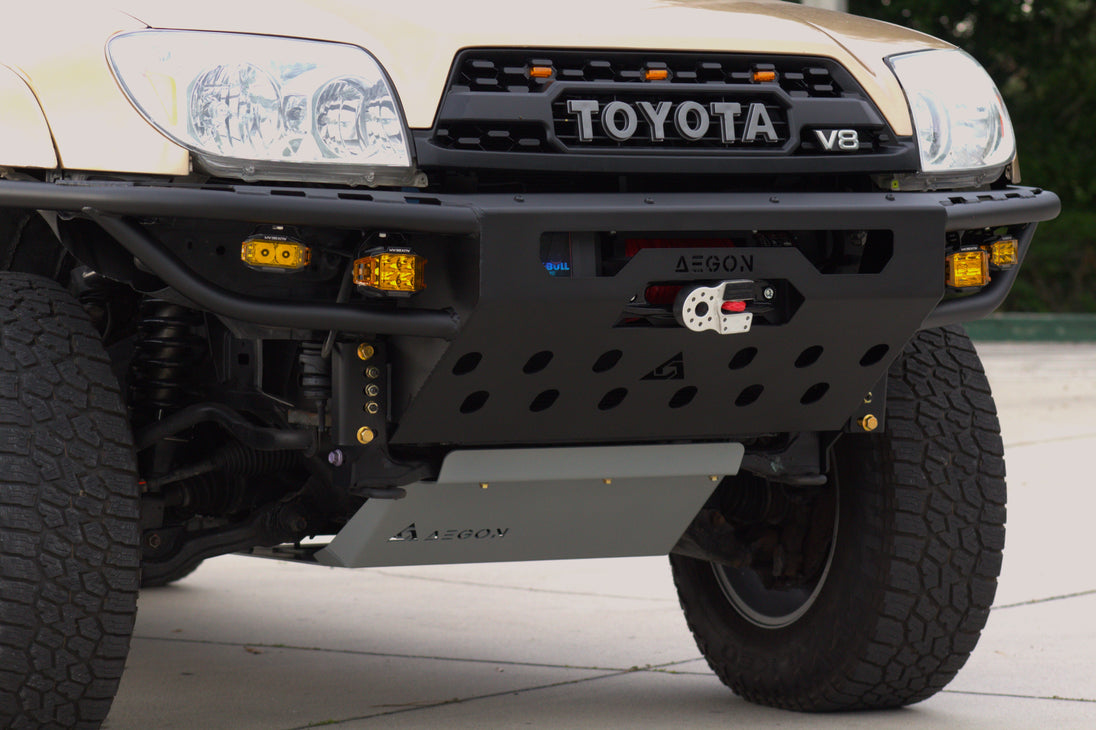 Close-up of the front bumper area of a Toyota V8 off-road vehicle, showing a black heavy-duty front bumper with integrated lights, a winch, and a silver skid plate on an outdoor paved surface.