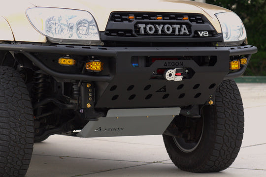 Close-up of the front bumper area of a Toyota V8 off-road vehicle, showing a black heavy-duty front bumper with integrated lights, a winch, and a silver skid plate on an outdoor paved surface.