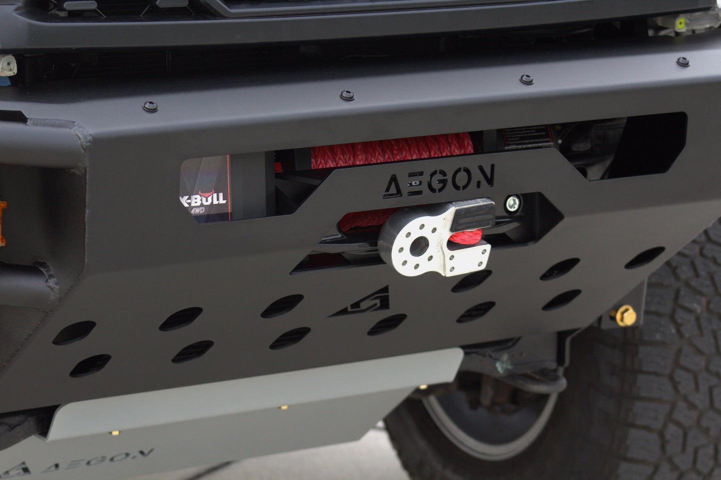 Close-up of a black metal front bumper with a winch mounted on it, featuring the AEGON brand name on the bumper and a winch housing with red and white components.