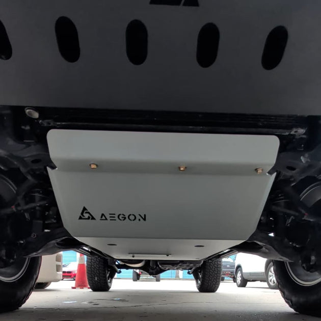 A close-up view from underneath a vehicle showing a white underbody shield with the AEGON logo printed on it, positioned above a gray concrete floor.